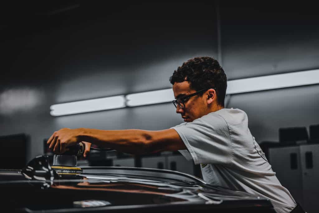 Person wearing glasses using a power tool to polish a car in a dimly lit garage.