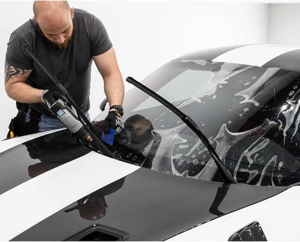 A person installs a windshield protection film on a black and white car using a power tool.