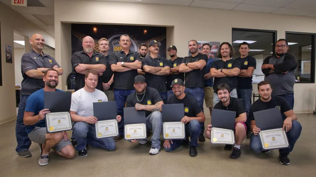 A group of men pose indoors, some standing with arms crossed, while others kneel in front holding certificates celebrating their expertise in windshield protection film.