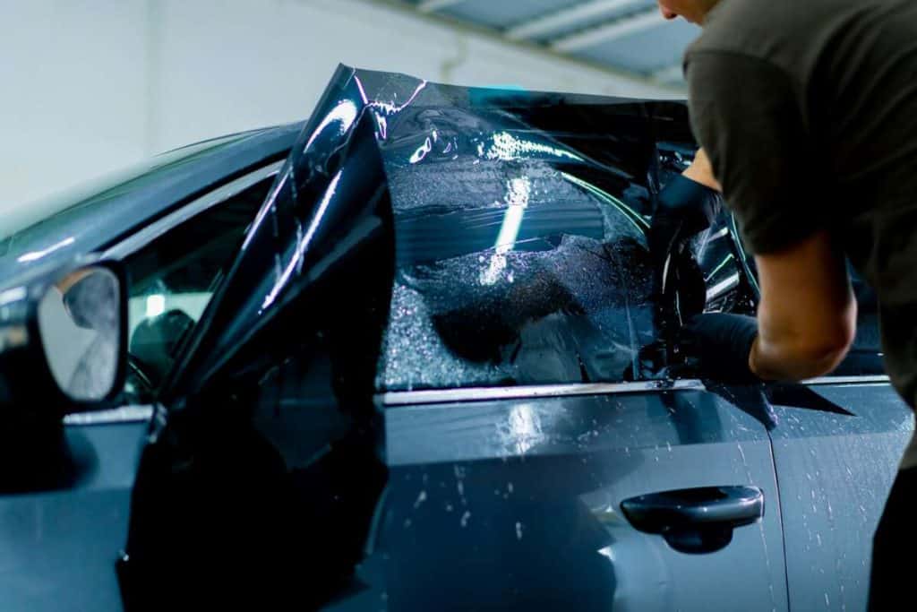 A person applies tinted windshield protection film to a car window inside a garage.