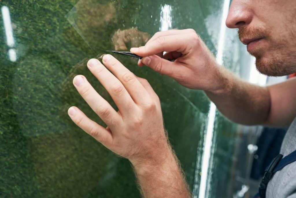 A person carefully applies a windshield protection film onto a glass surface using a small tool, focusing on precision and alignment.