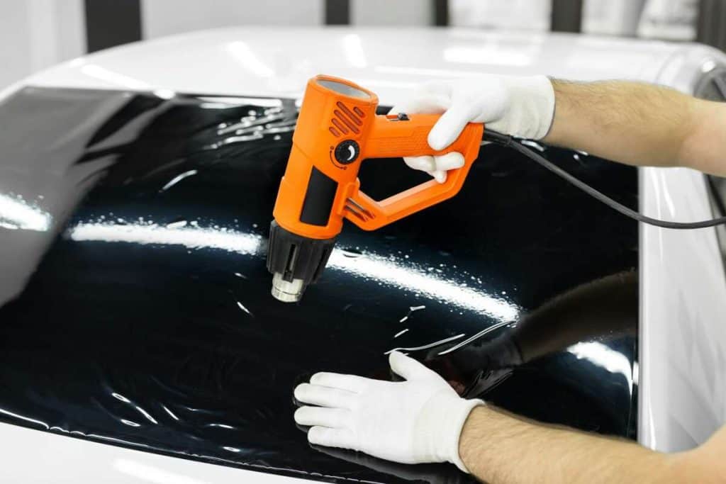 A person in white gloves uses an orange heat gun to expertly apply or remove tint film on a car window, ensuring optimal windshield protection.