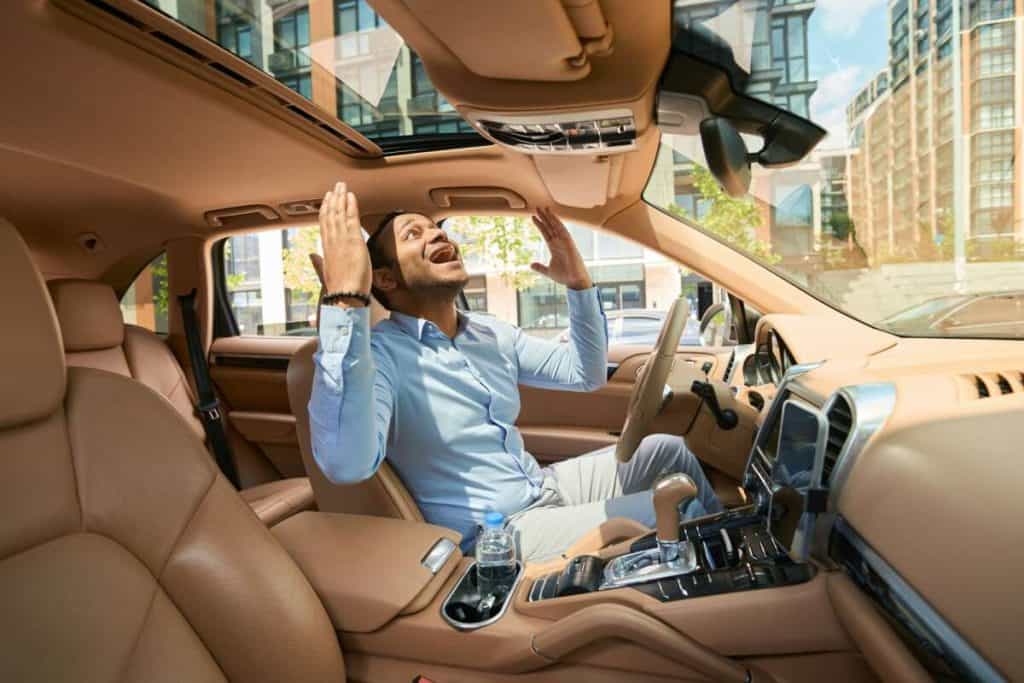 A person in a car, featuring a sleek tan interior and ceramic window tint, gestures upwards in frustration. Sunlight filters softly through the tinted windows as city buildings stand visible outside.