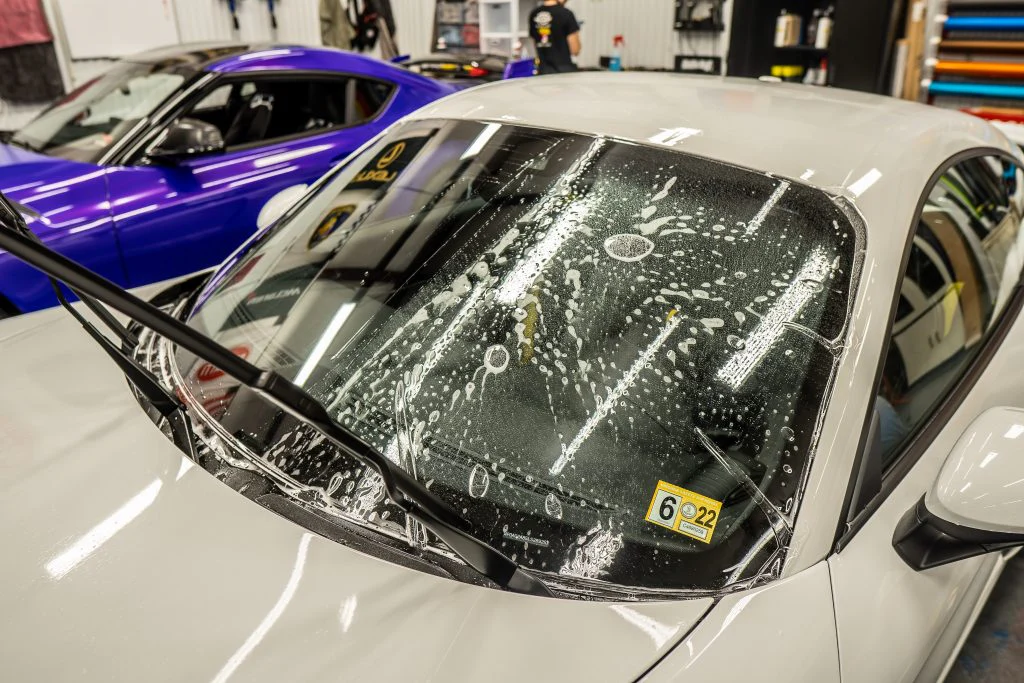 Close-up of a white car with soapy water on the windshield, highlighting its new windshield protection film. A blue car is seen next to it in the background, inside a garage.