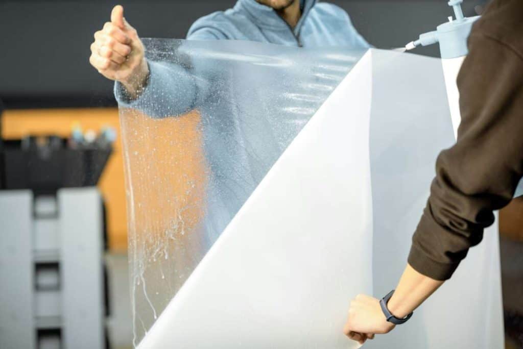 Two people carefully manage a large transparent plastic sheet, as if it were a windshield protection film.