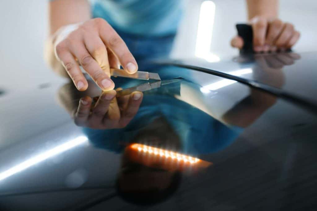 A person meticulously uses a tool to inspect or repair the car hood, ensuring every detail is perfect before applying the sleek finish of ceramic window tint that will enhance both style and UV protection.