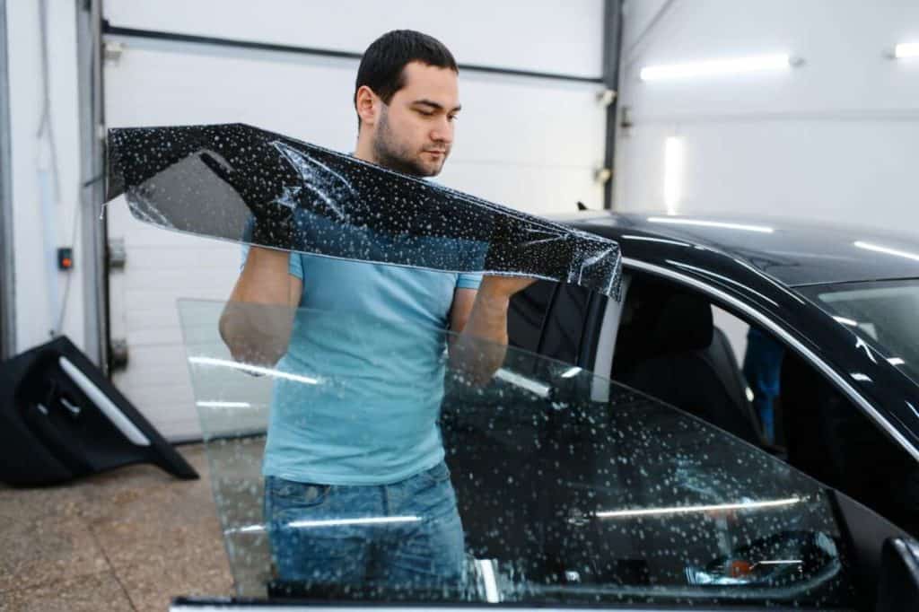 In the garage, a person applies a tinted film to the car window, carefully aligning it for optimal windshield protection.