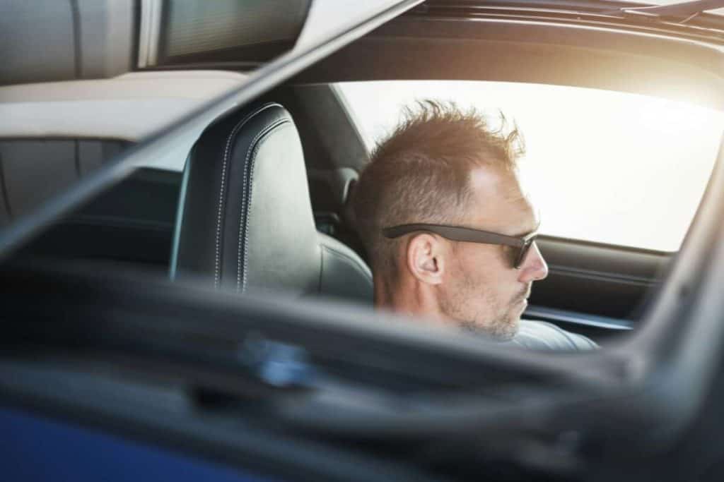 A person with sunglasses and a spiky hairstyle is sitting in the driver's seat of a car, their features subtly obscured by the sleek ceramic window tint viewed through the open window.