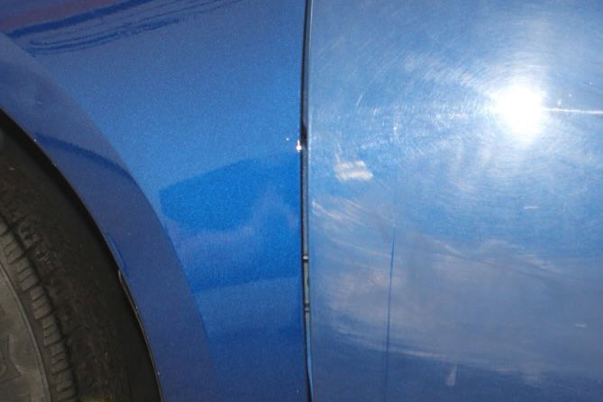 Close-up of a blue car showing a small dent and a paint scratch near the wheel arch.